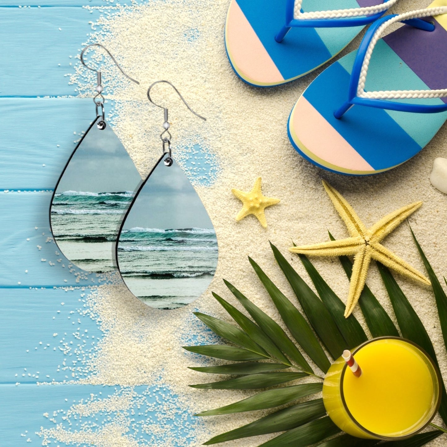 Teardrop-shaped earrings with beach scene design, blue flip-flops, starfish, palm leaves, and a yellow drink on a sandy surface.