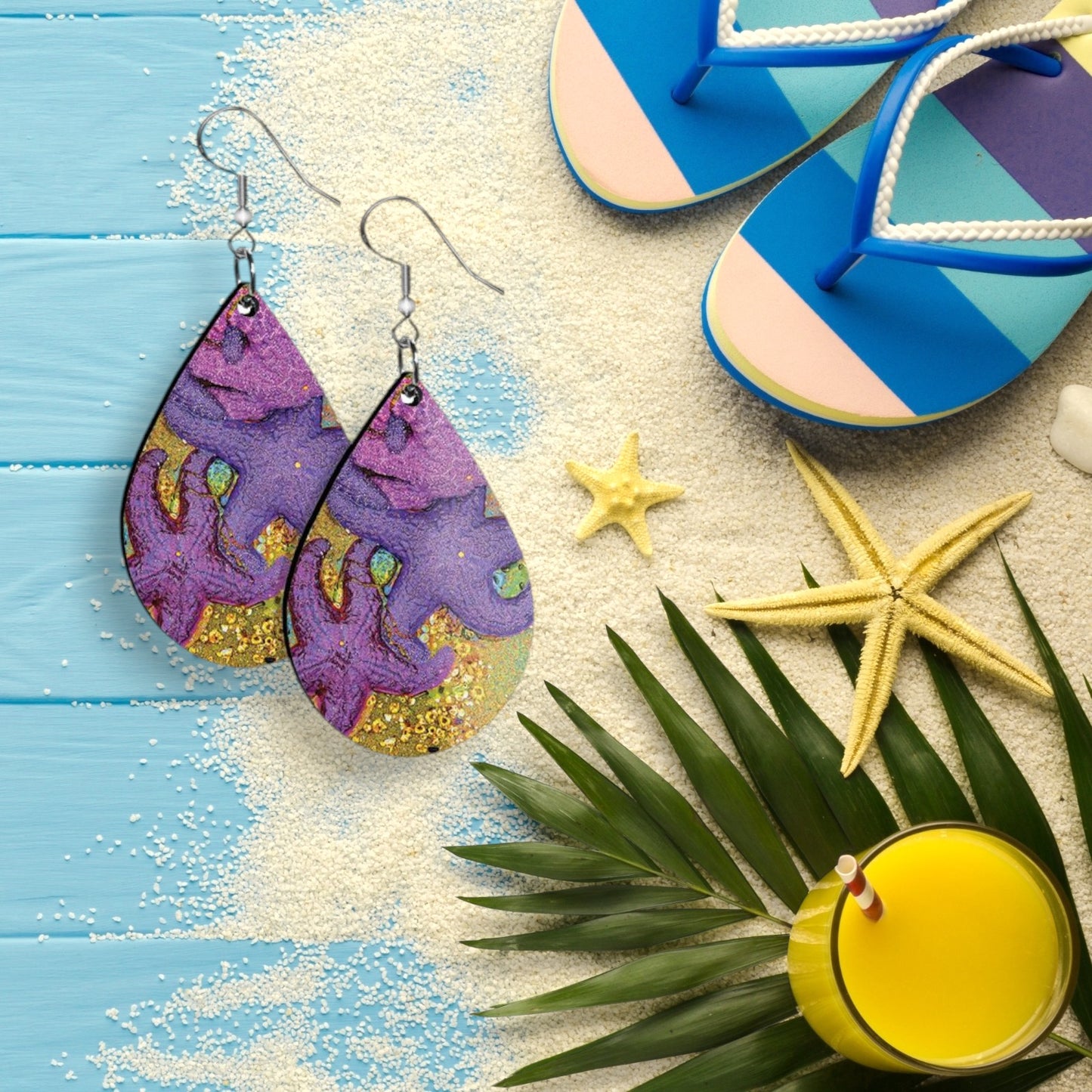 Colorful earrings on a sandy surface with flip-flops, starfish, and a palm leaf.