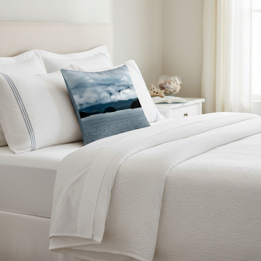 A styled bed with white bedding and pillows, featuring a square decorative throw pillow printed with a scenic landscape of blue water and cloudy sky.