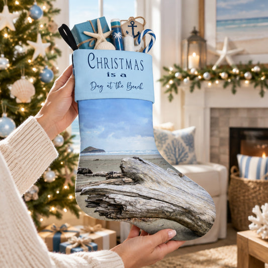 A person holds a Christmas stocking in a cozy living room setting. The stocking features a printed beach scene with driftwood and the text 'CHRISTMAS is a Day at the Beach' on a light blue band.