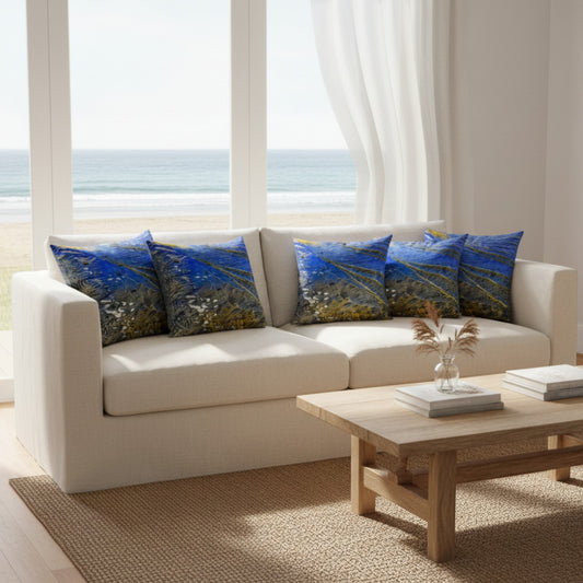 A cream-colored fabric sofa in a bright, modern living room setting with a wooden coffee table and a large window overlooking the ocean. The sofa is styled with four decorative throw pillows featuring a textured blue and yellow abstract landscape pattern.