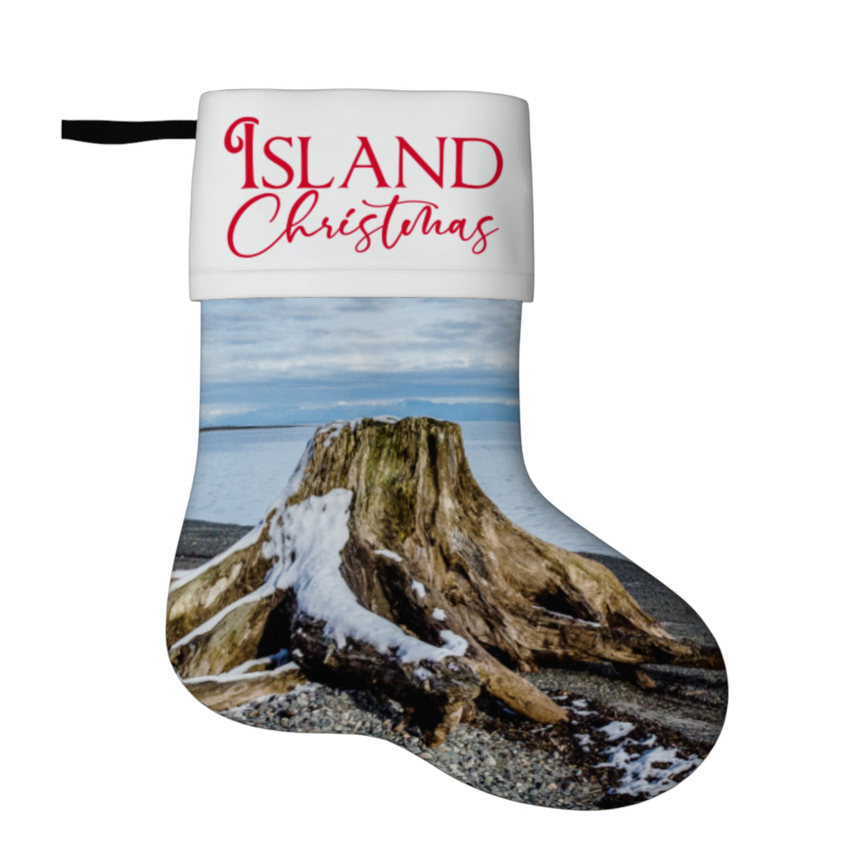 A Christmas stocking with a white cuff printed with the words 'ISLAND Christmas' in red script, and the body featuring a photographic print of a driftwood log on a rocky beach with snow and water under a cloudy sky.