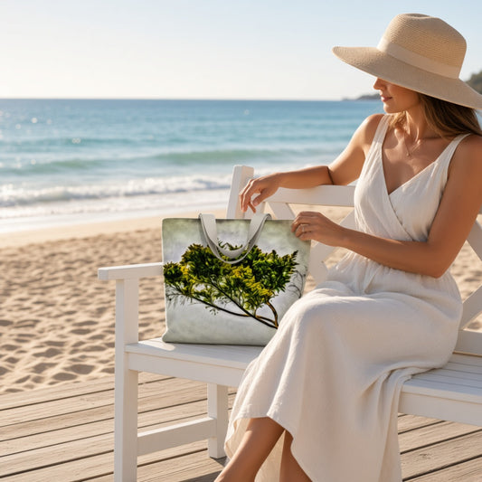 A woman in a white dress and straw hat sits on a white beach chair at the beach, holding a tote bag with a green tree print and silver handles.