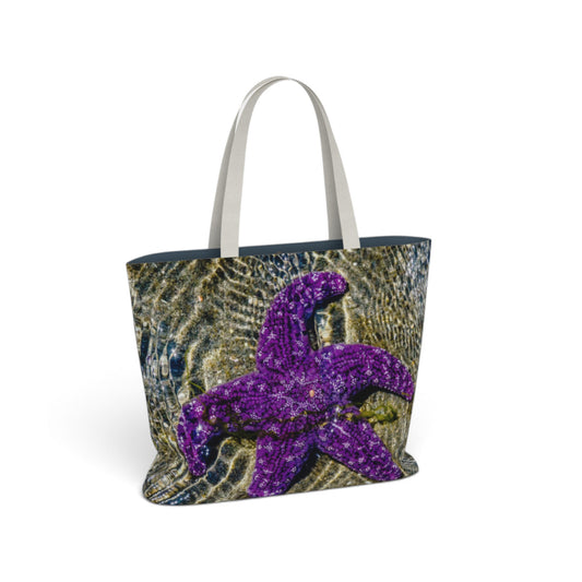 A tote bag featuring a printed design of a purple starfish over a blue and gold underwater wave pattern, with light gray shoulder straps, shown on a white background.