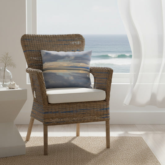 A woven outdoor armchair with a striped blue and brown wicker pattern, a white seat cushion, and a matching blue and beige striped throw pillow, set in a bright room with a window overlooking the ocean.