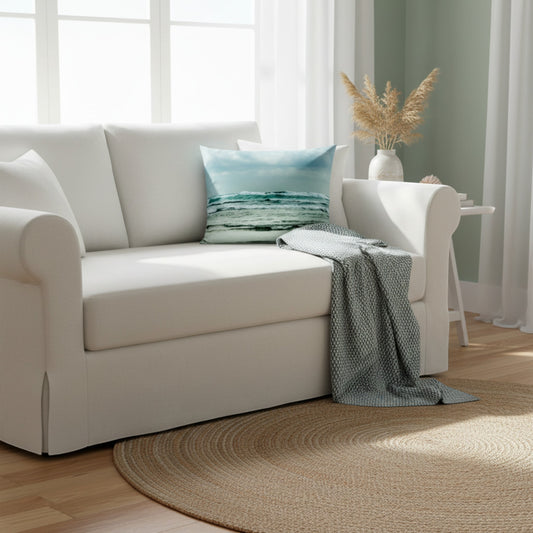A white upholstered loveseat in a bright living room, styled with a blue ocean-print throw pillow and a draped gray patterned blanket, set on a round woven rug.