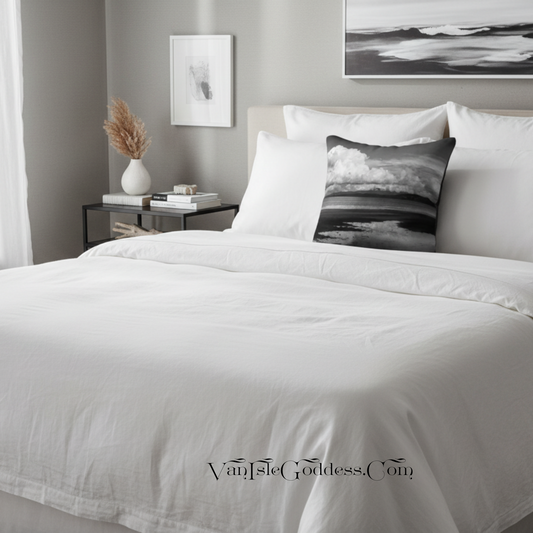 A neatly made bed with white bedding and a black-and-white landscape throw pillow in a styled bedroom. A side table with books and a vase, wall art, and a window curtain are visible in the background.