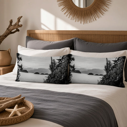 A styled bedroom scene with a bed made up with white and dark gray bedding and two long rectangular pillows featuring a black-and-white coastal landscape print with trees and distant hills. A wicker headboard, sunburst wall mirror, and decorative tray on the bed are visible.
