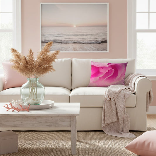 A framed wall art print depicting a soft ocean sunset hangs above a cream sofa in a bright living room. The room includes a pink pillow, a beige throw blanket, a glass vase with dried grass, and a light wood coffee table.