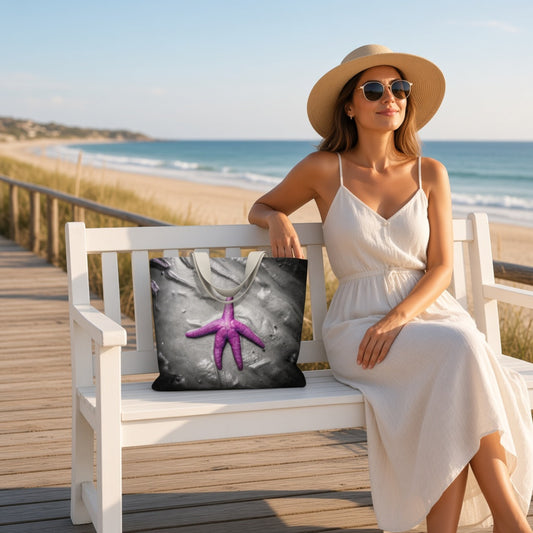 A woman in a white sundress and straw hat sits on a white wooden beach boardwalk bench, holding a tote bag printed with a purple starfish on a gray rock background. The beach and ocean are visible in the background.