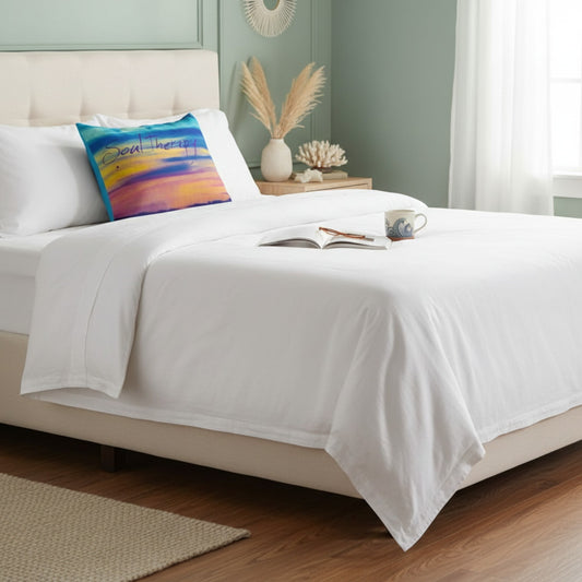 A neatly made bed with crisp white bedding and a colorful abstract throw pillow in a softly styled bedroom with pale green walls, wood flooring, and bedside decor.