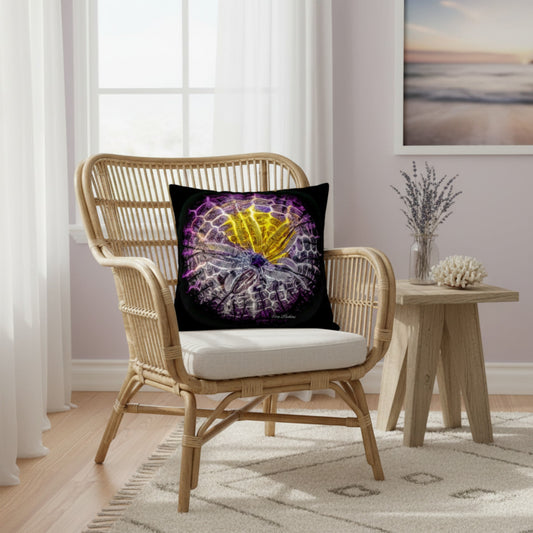A square throw pillow with a vibrant purple and yellow spiderweb design on a black background, styled on a light wood rattan armchair in a bright, modern room.
