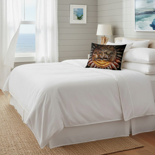 A neatly made bed with crisp white bedding and a decorative black sun-face throw pillow, set in a bright bedroom with white curtains, a window overlooking the ocean, and light gray paneled walls.