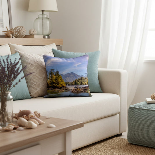 A square throw pillow featuring a scenic mountain lake landscape print, styled on a cream-colored sofa in a bright, modern living room.