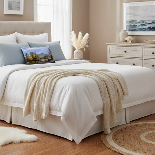 A neatly made bed in a softly styled bedroom, dressed in white bedding with a beige knit throw draped across the foot. Light blue pillows and a small landscape accent pillow are visible, with a dresser, framed wall art, and wood flooring in the background.