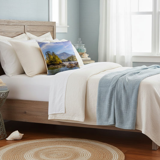 A styled bedroom scene showing a bed with white bedding, a light blue throw blanket, and a square decorative pillow featuring a scenic landscape print of mountains and trees.