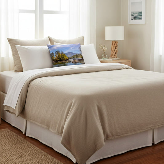 A neatly made bed in a bright bedroom setting, dressed with a light beige comforter, white pillows, and a decorative square pillow featuring a colorful landscape print.