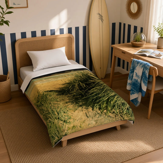 A single bed in a styled bedroom setting, featuring a duvet cover printed with a beach grass landscape design in green and beige tones. The room includes a wooden desk, a surfboard leaning against the wall, and a striped accent wall.