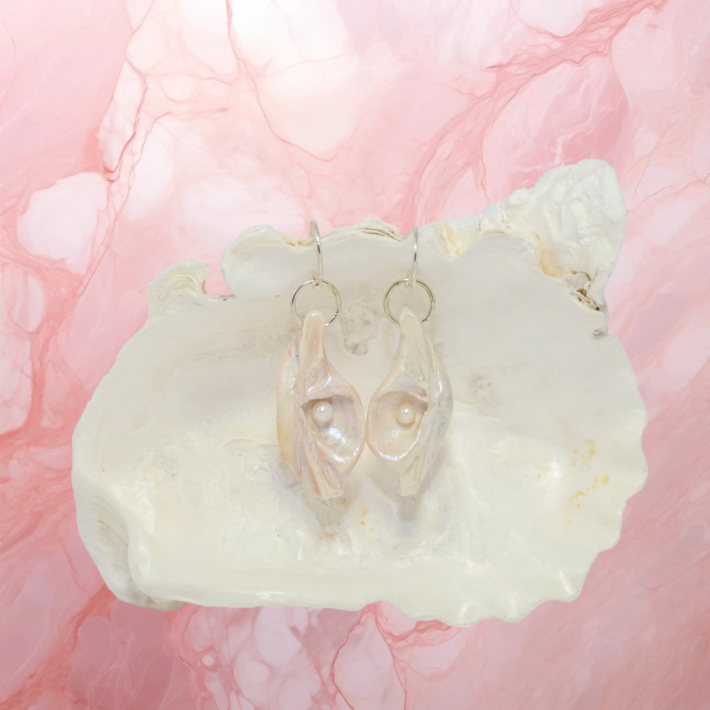 Victoria natural seashell earrings with real baby freshwater pearls. The earring are in front of a larger seashell with a marbled pink background.