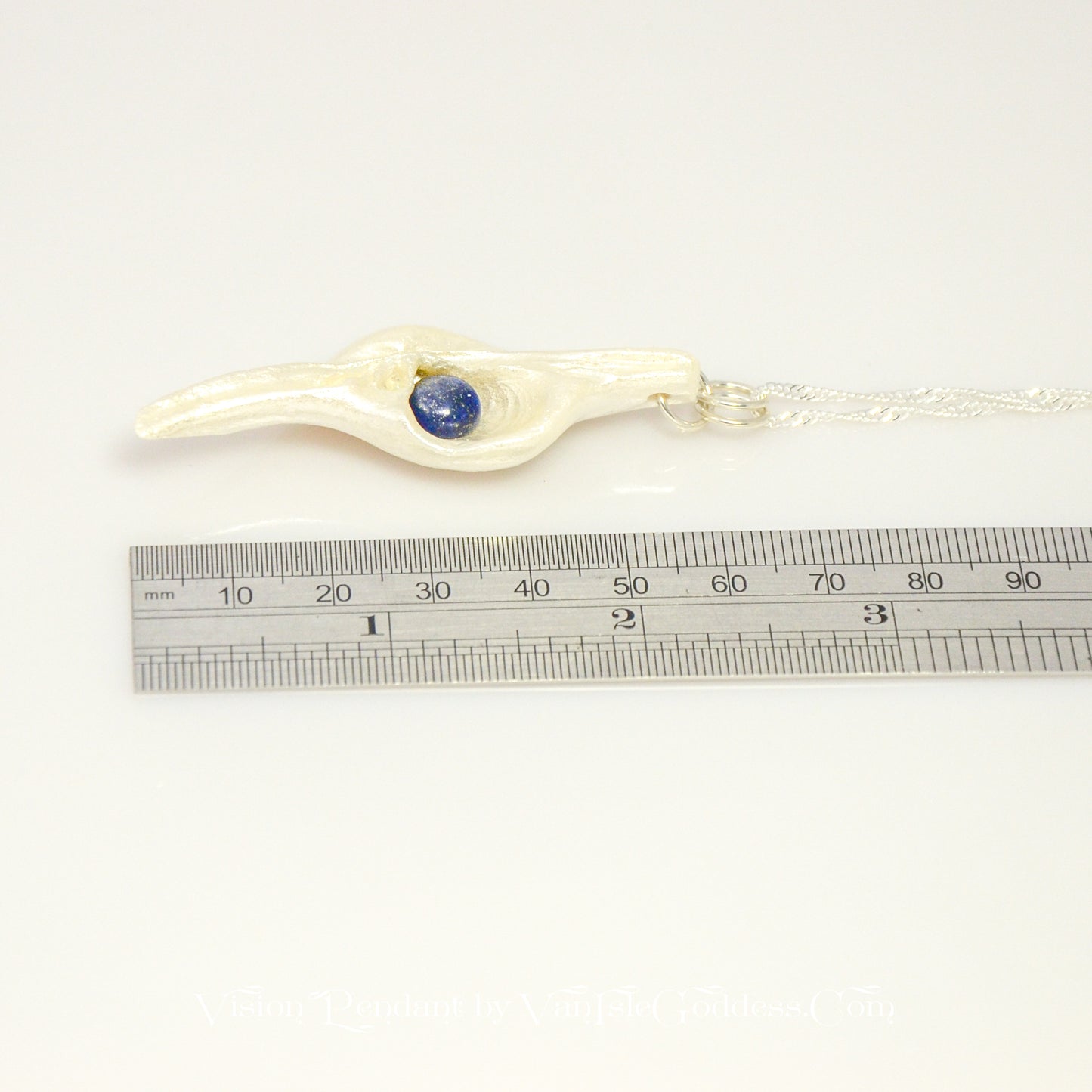 Vision is a natural seashell pendant with a Lapis Lazuli gemstone. The pendant lays along side a ruler so the viewer can see the length of the pendant.