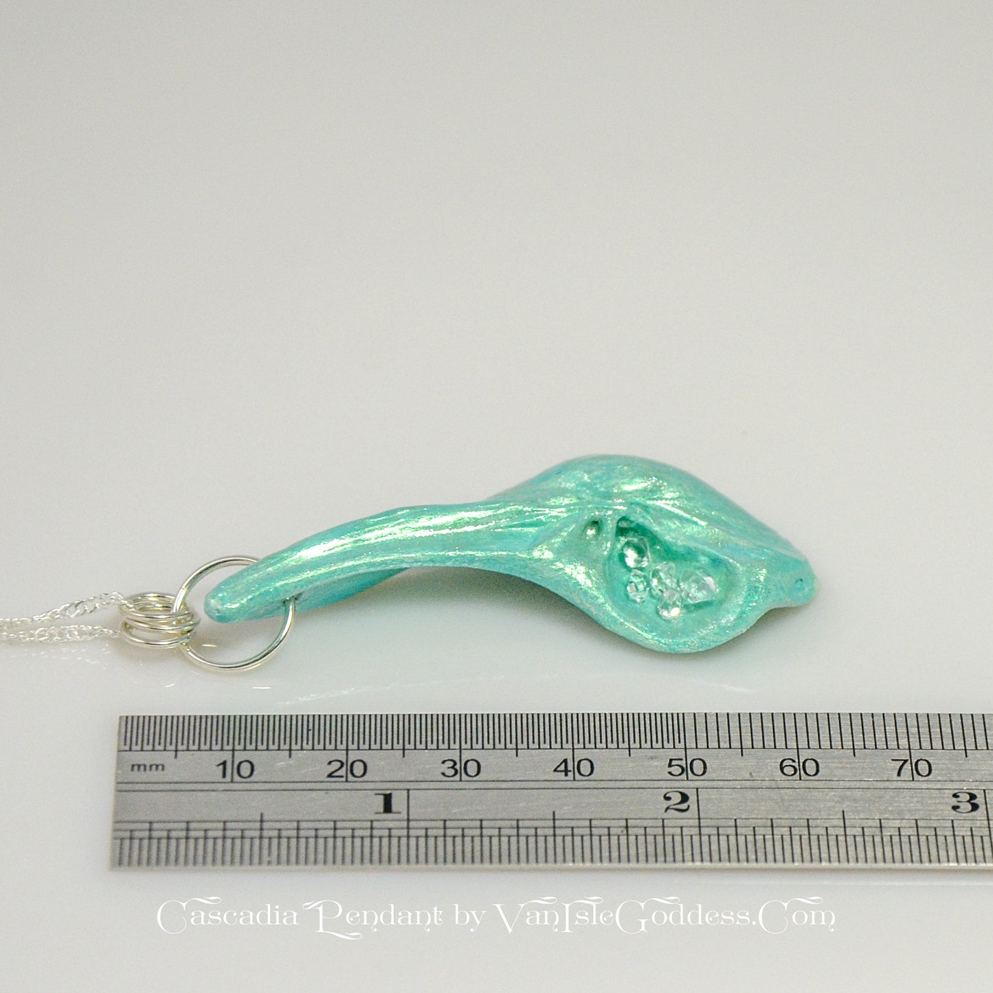 The Cascadia Pendant created with a natural seashell from the beaches of Vancouver Island. The seashell is turquoise and has high quality herkimer diamonds. The pendant is shown on its side with a ruler so the view can see the length of the pendant. The print on the bottom of the image says: Cascadia Pendant by Van Isle Goddess dot com