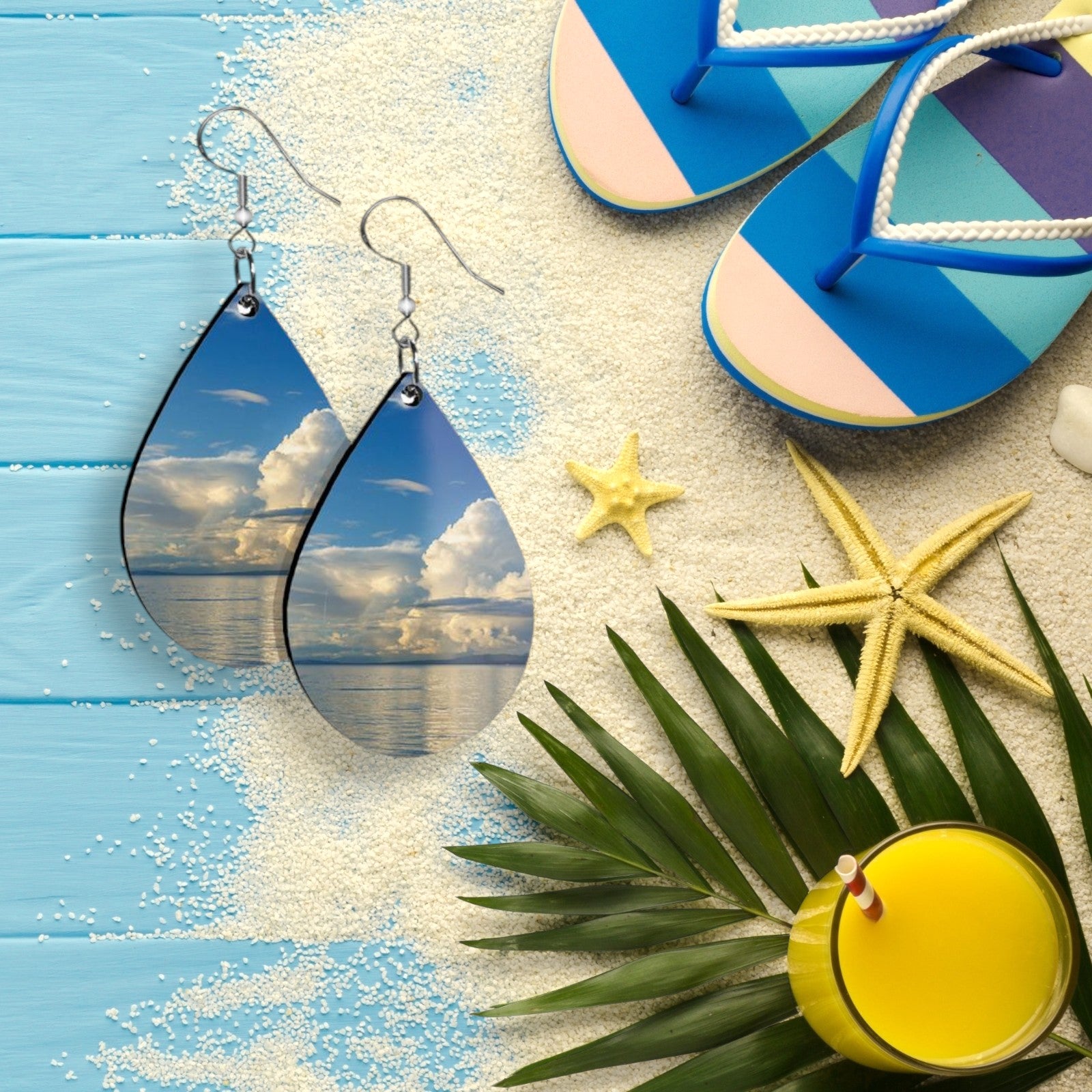 Teardrop-shaped earrings with beach scene design, placed on a sandy surface with flip-flops, starfish, and a palm leaf.