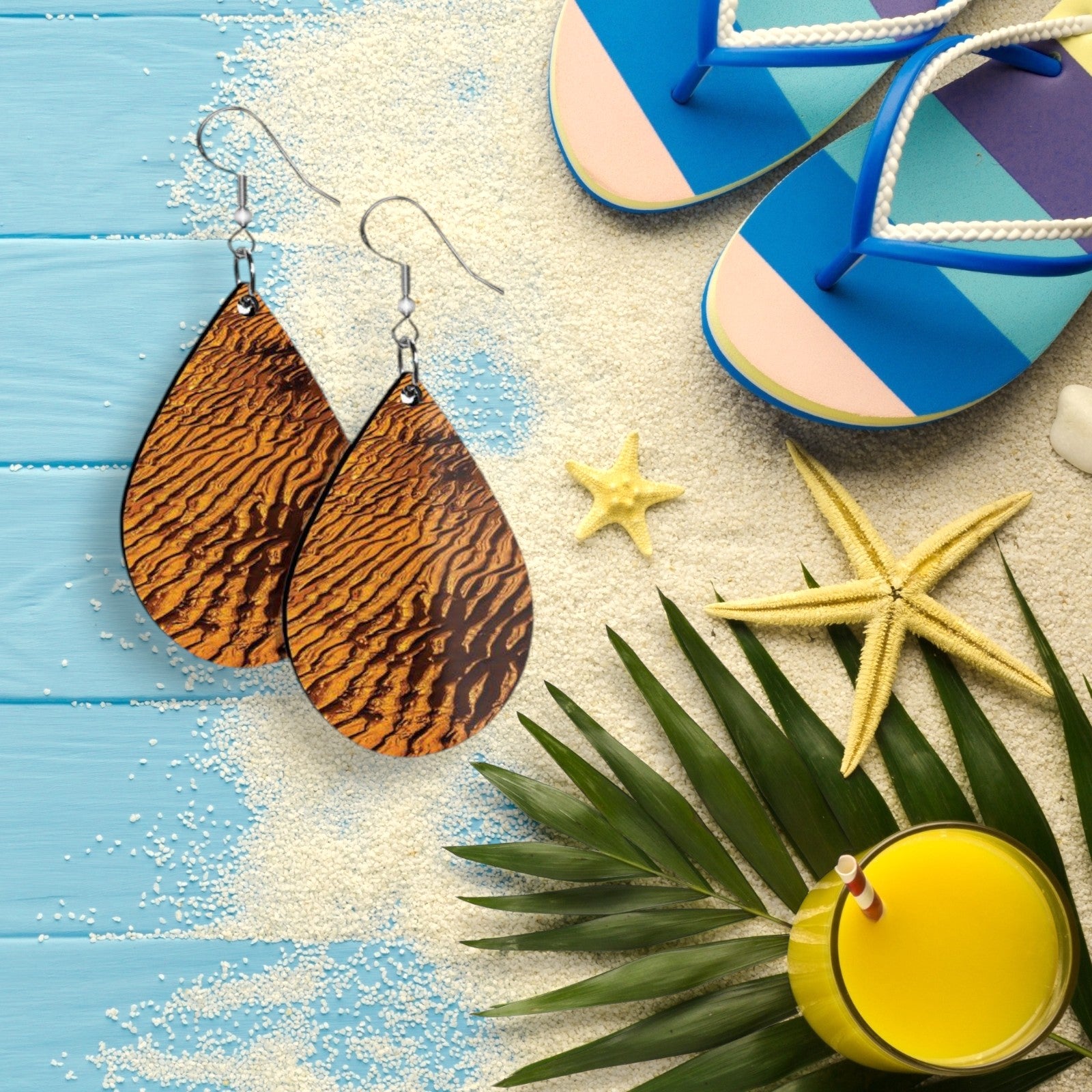 Tropical-themed setting with sand-striped earrings, starfish, flip-flops, and a drink on a blue wooden surface.