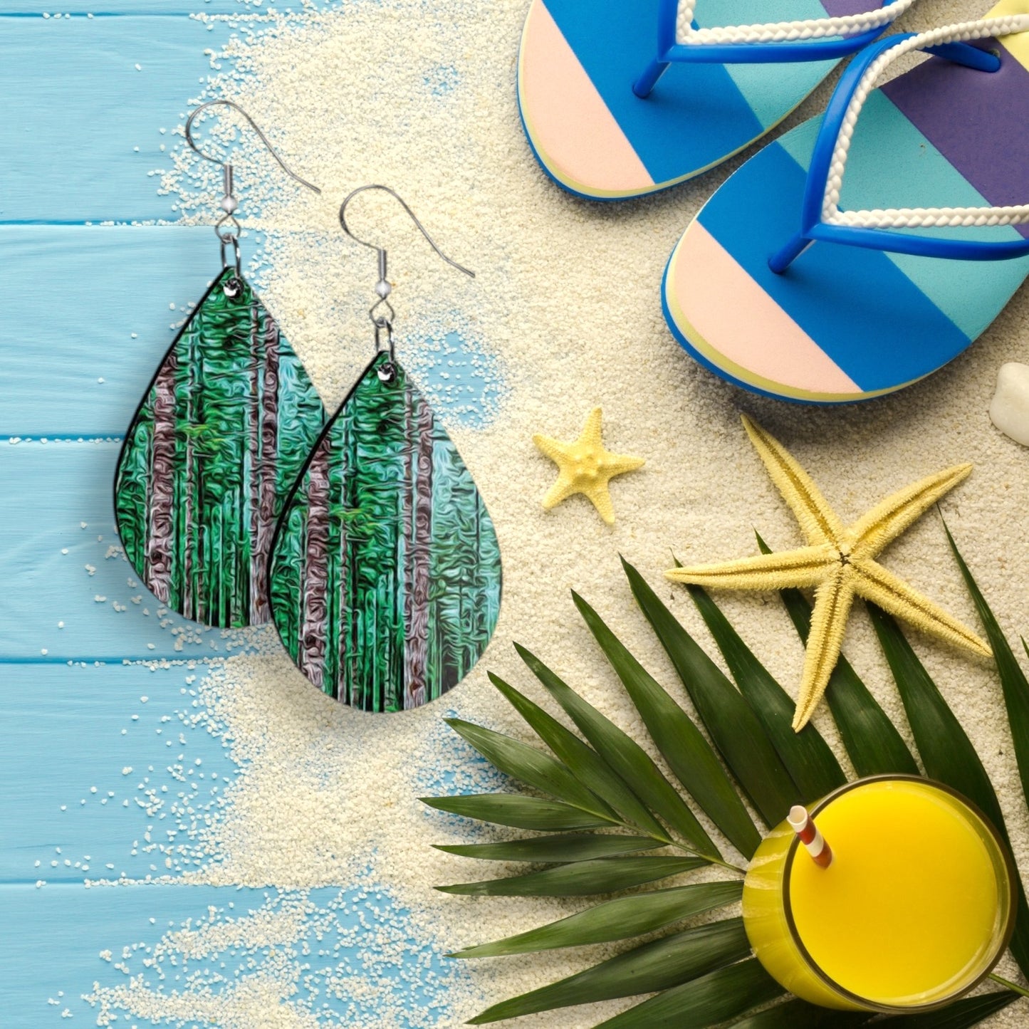 Teardrop-shaped earrings with a forest design on a beach-themed background with flip-flops, starfish, and a palm leaf.