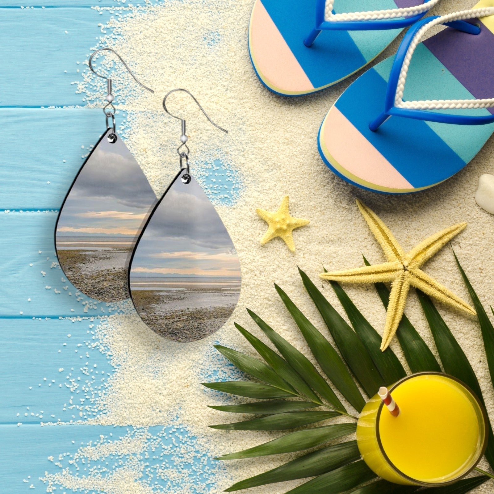 Teardrop-shaped earrings with a beach scene, blue flip-flops, starfish, palm leaves, and a yellow drink on a wooden surface.