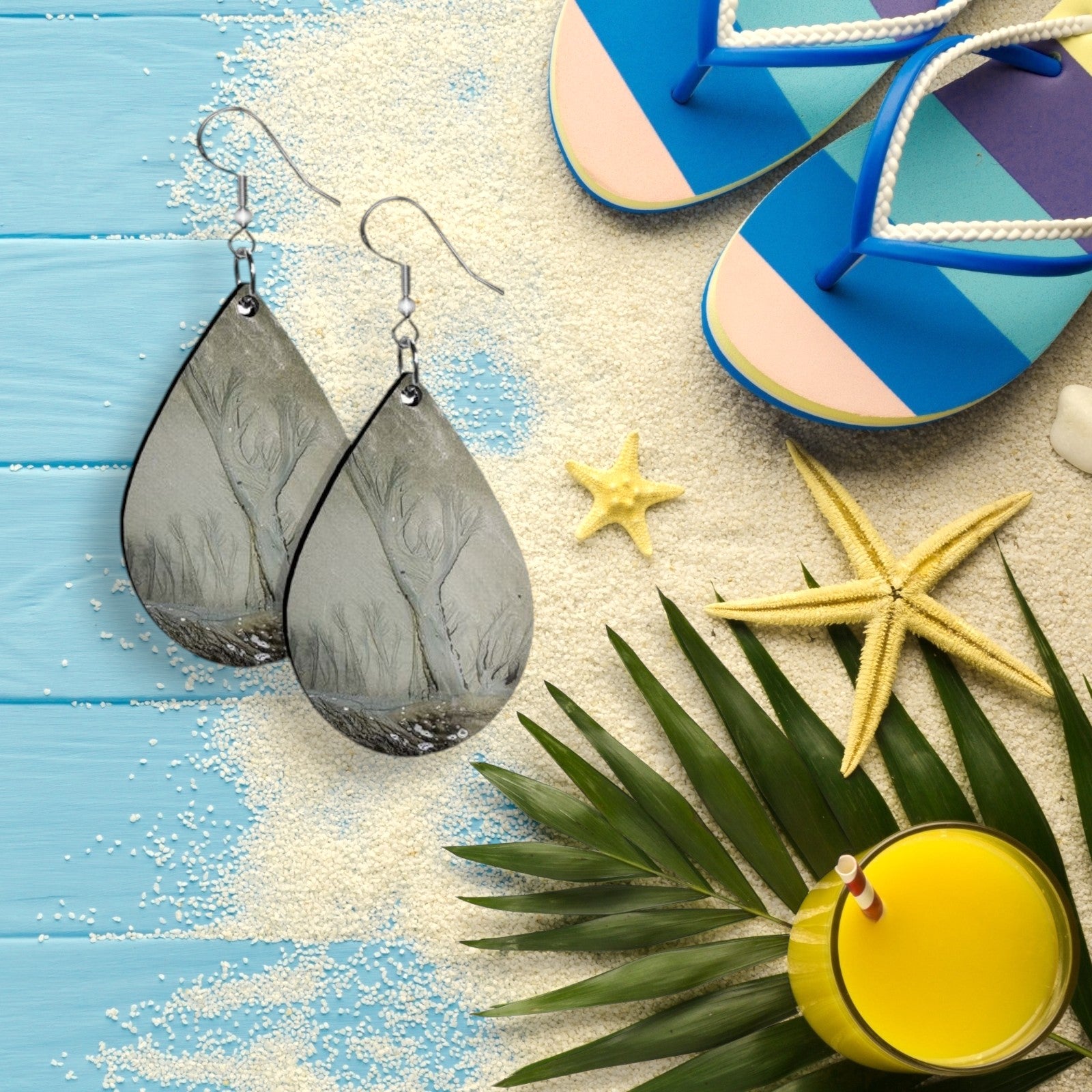 Sand teardrop earrings on a sandy surface with flip-flops, starfish, and a palm leaf.