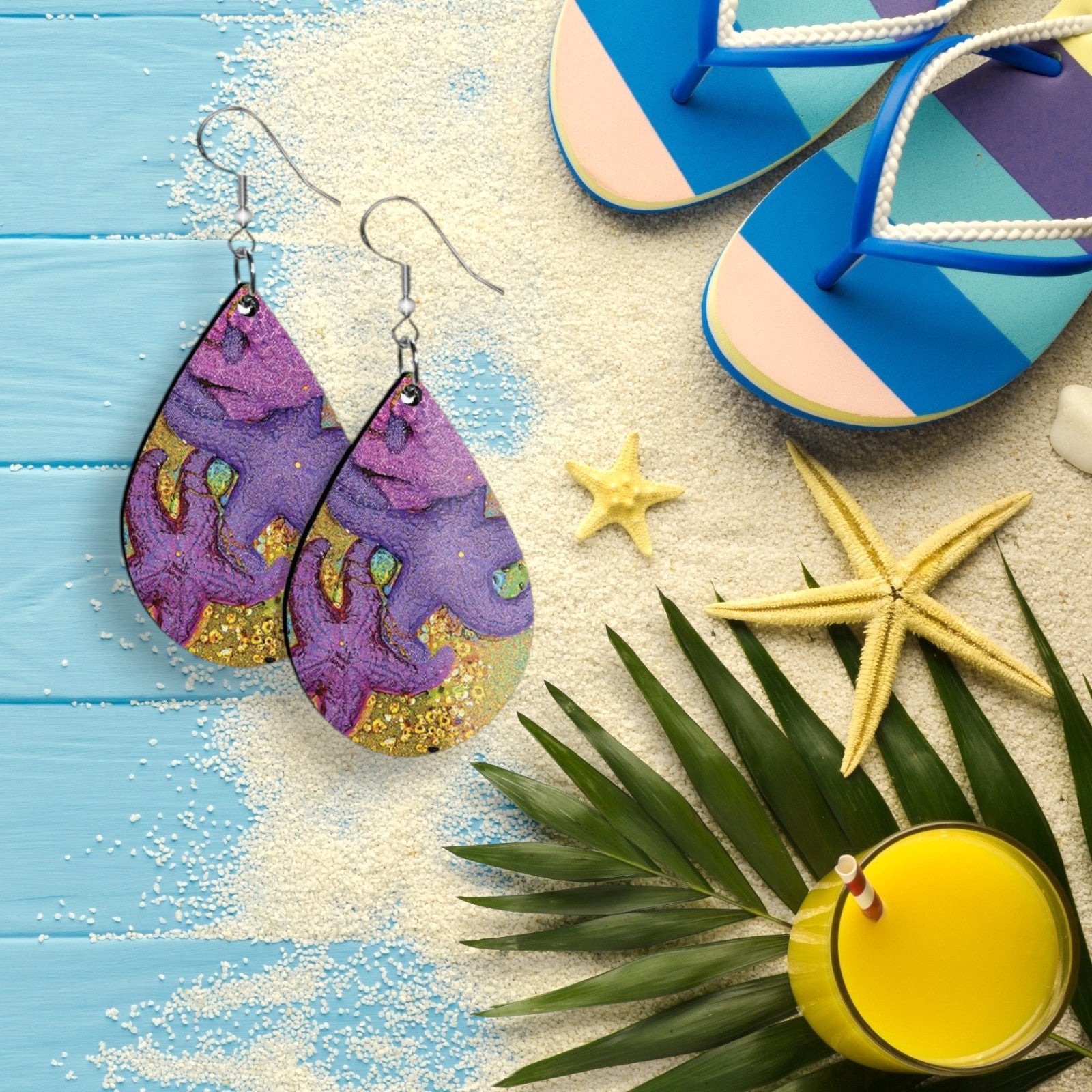 Colorful earrings on a sandy surface with flip-flops, starfish, and a palm leaf.