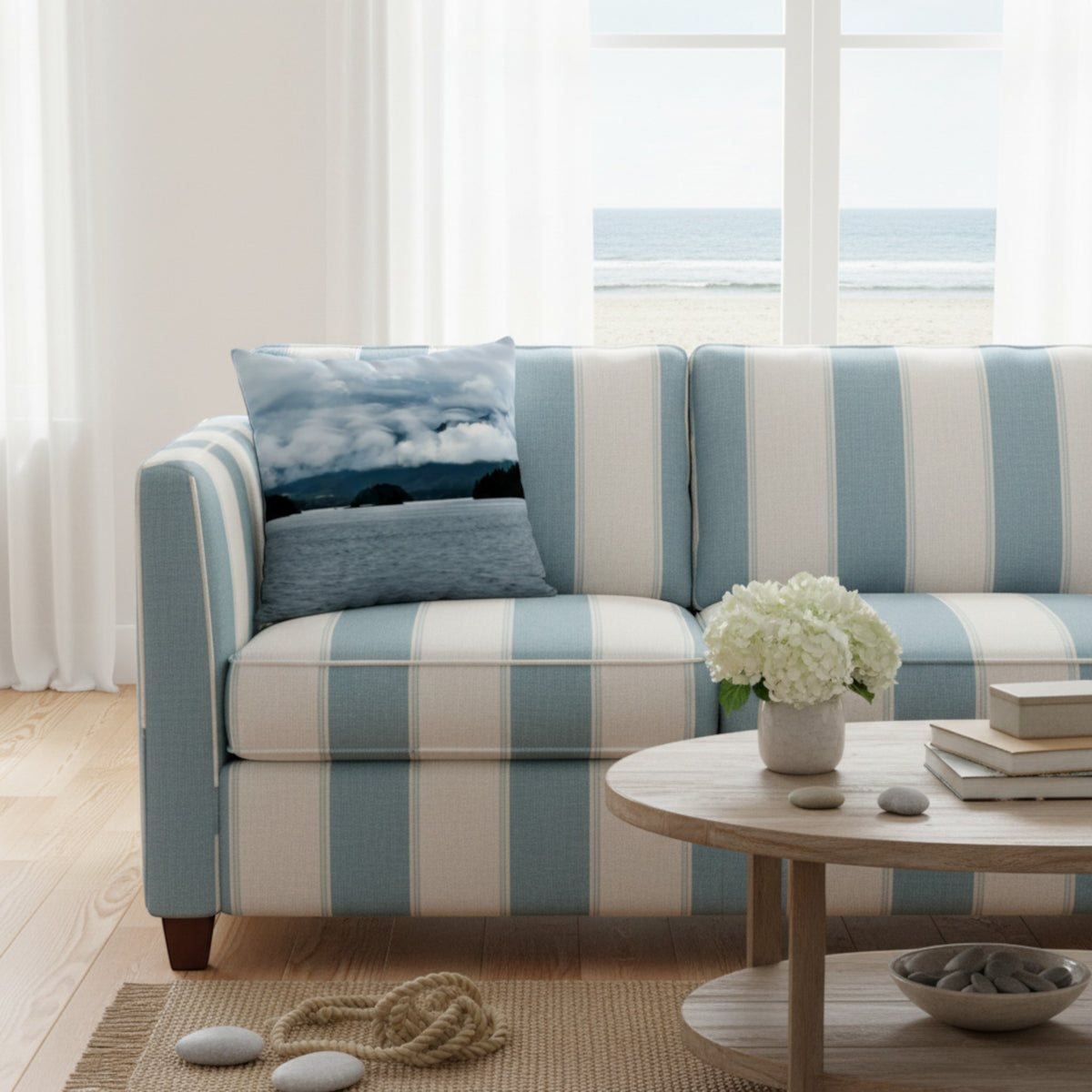 A blue and cream striped sofa in a bright, modern living room with a view of the ocean through large windows. A matching blue and cream striped throw pillow with a landscape print rests on the sofa, alongside a round wooden coffee table with white flowers.
