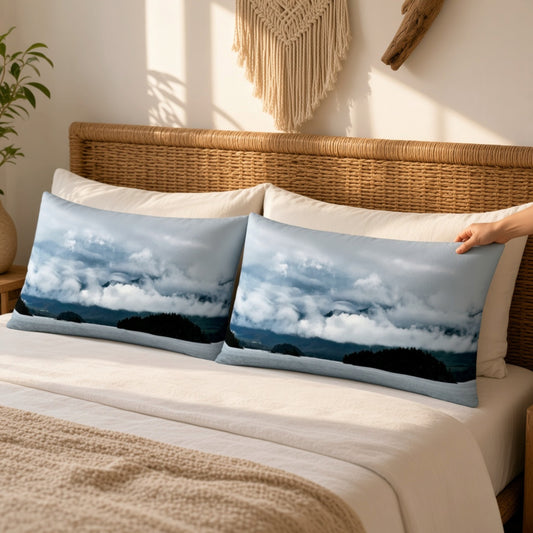 Two rectangular throw pillows with a scenic landscape print of clouds and mountains, styled on a bed with a wicker headboard in a warm, sunlit room.