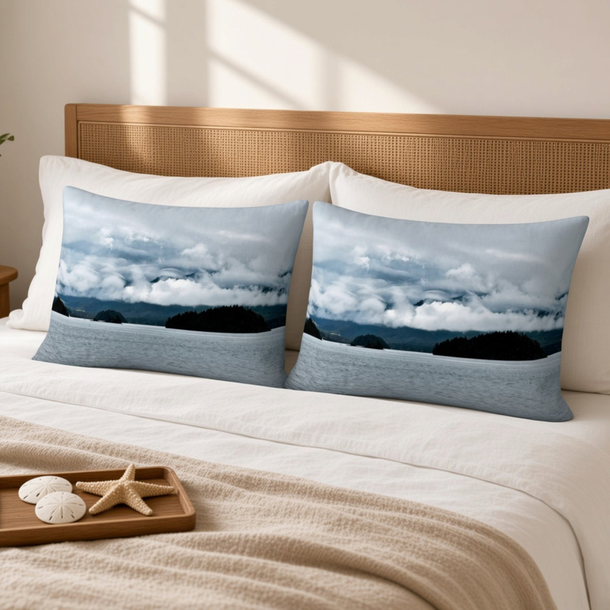 Two rectangular decorative pillows with a blue-gray seascape print are styled on a bed with white pillows, a beige blanket, and a wooden headboard in a softly lit room.
