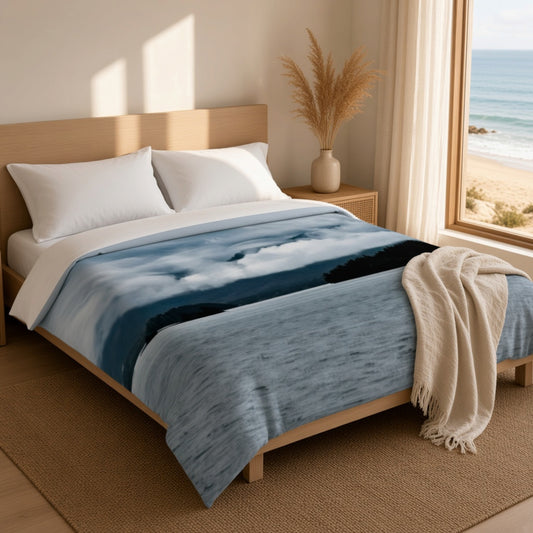 A light wood bed frame dressed with white pillows and a blue-and-white cloud-print blanket, styled in a bright bedroom with a window overlooking the ocean and a beige throw draped over the foot of the bed.