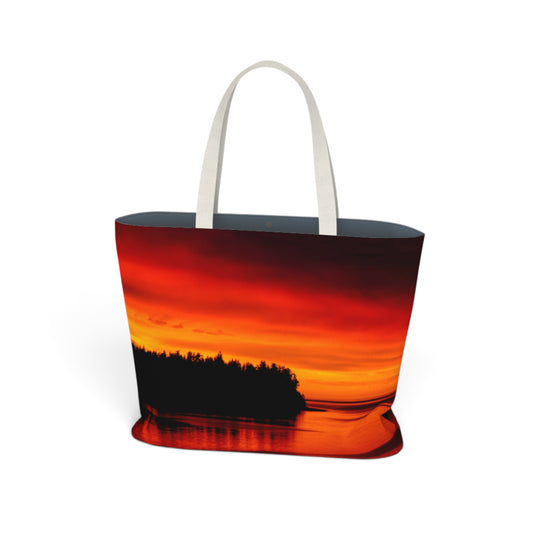 A tote bag featuring a vibrant sunset landscape print with red, orange, and yellow tones over a dark tree silhouette, complete with white shoulder straps, shown on a plain white background.
