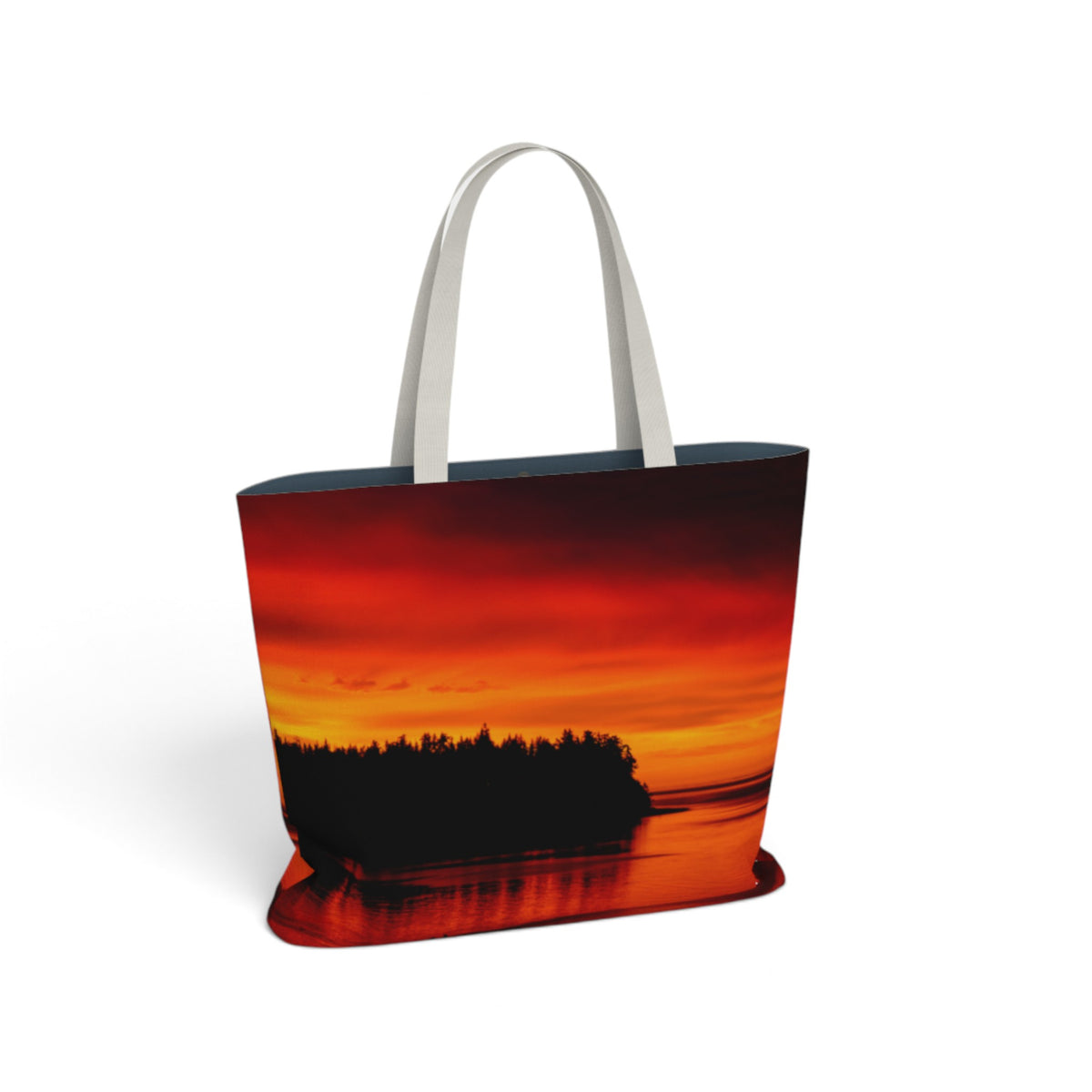 A tote bag featuring a vibrant sunset landscape print with red, orange, and yellow tones over a dark tree silhouette, complete with light gray shoulder straps, shown on a plain white background.