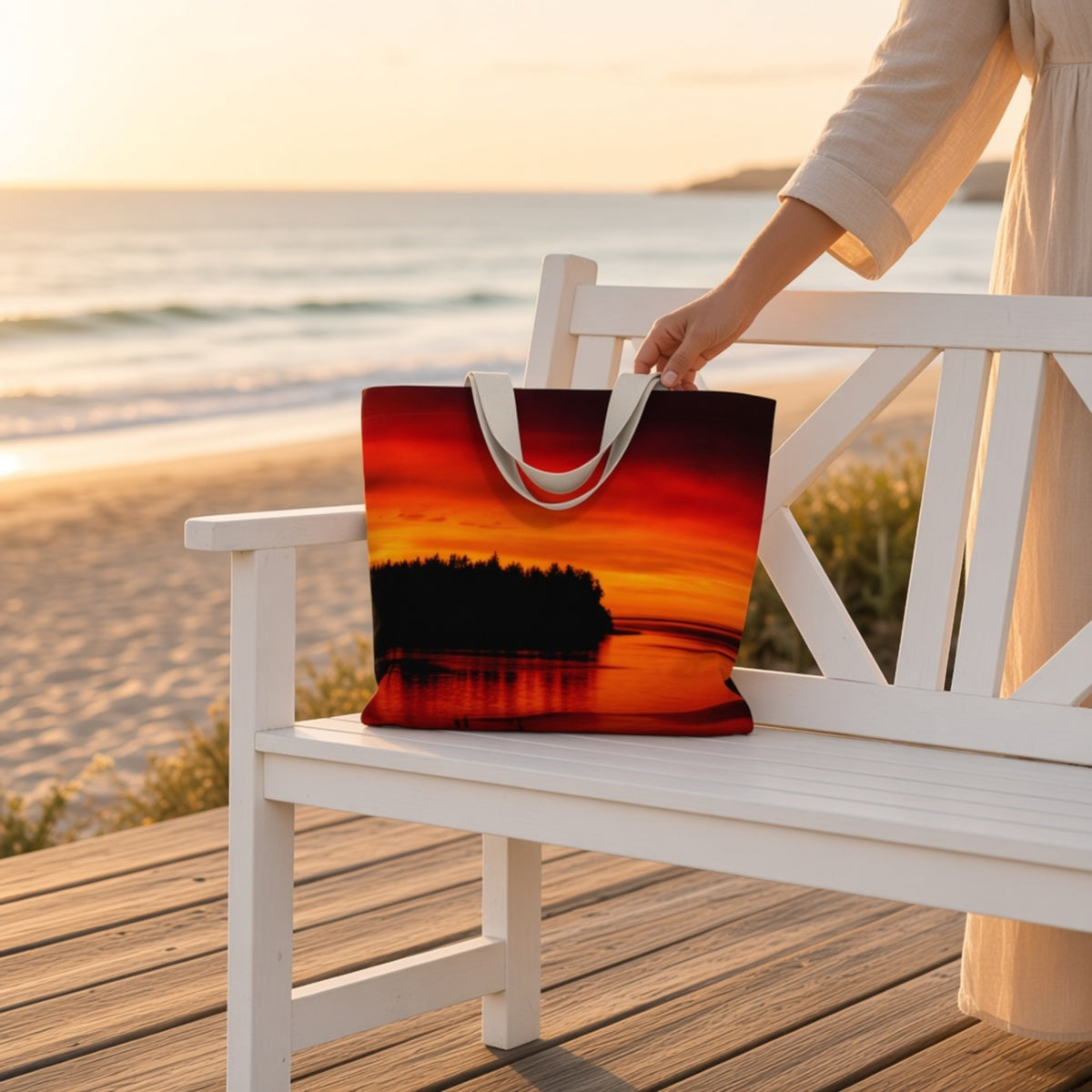 A tote bag with a sunset landscape print sits on a white wooden bench at the beach, with a person in a cream dress holding one handle.