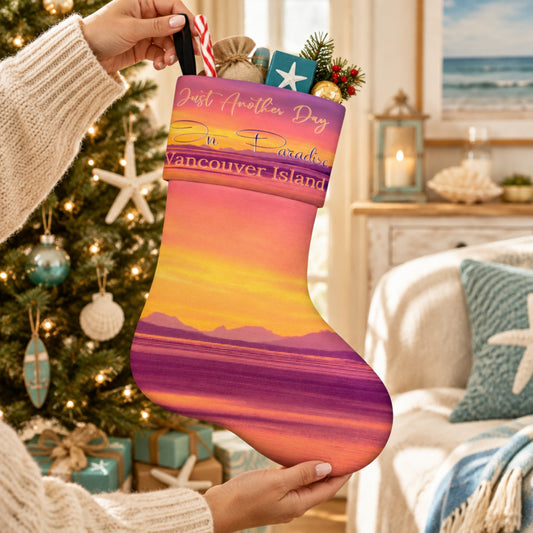A person holds a Christmas stocking printed with a sunset beach scene and the text 'Just Another Day In Vancouver Island.' The stocking is shown in a cozy living room with a decorated Christmas tree, candles, and a couch in the background.