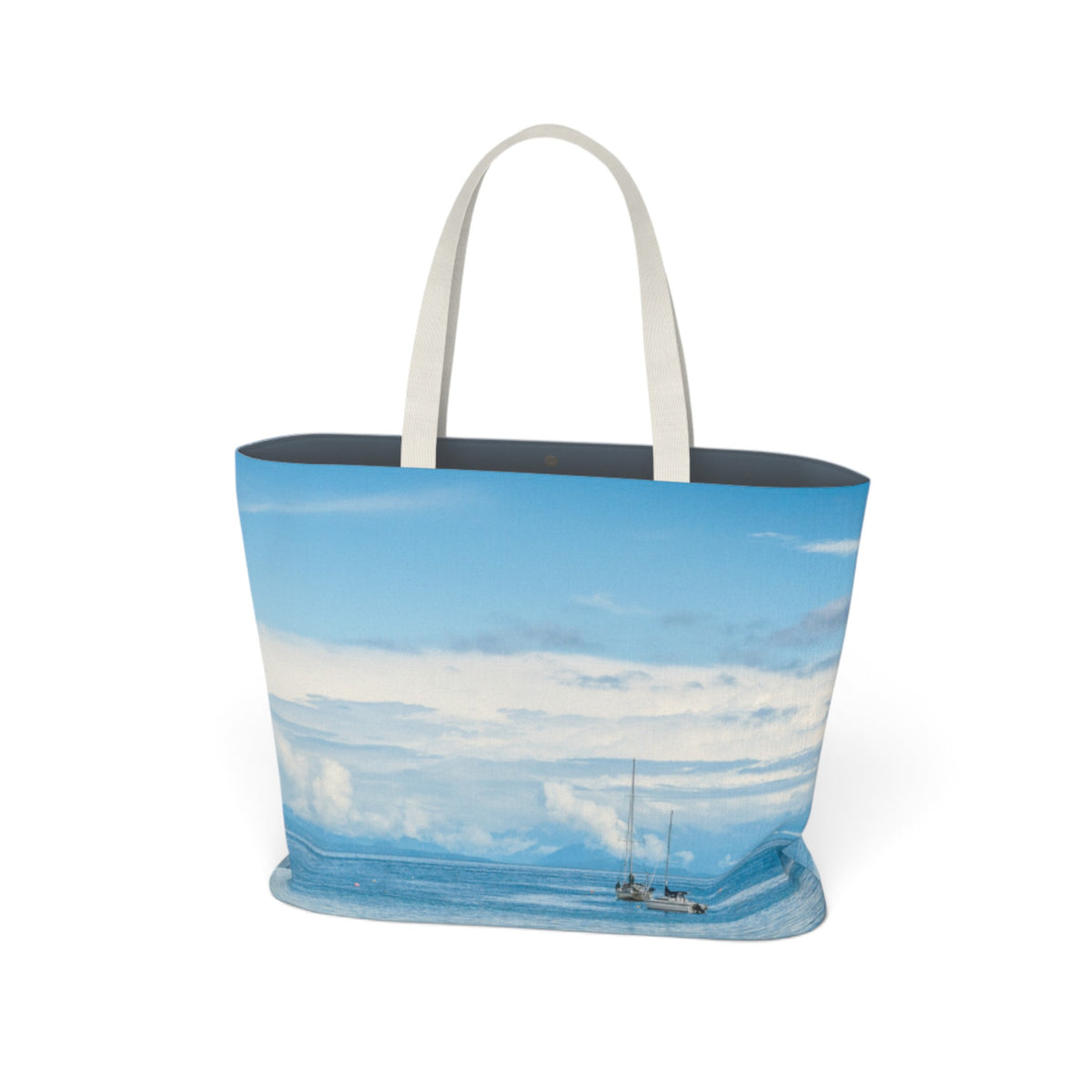 A tote bag featuring a scenic print of a sailboat on a blue ocean under a cloudy sky, with white fabric shoulder straps, shown against a plain white background.