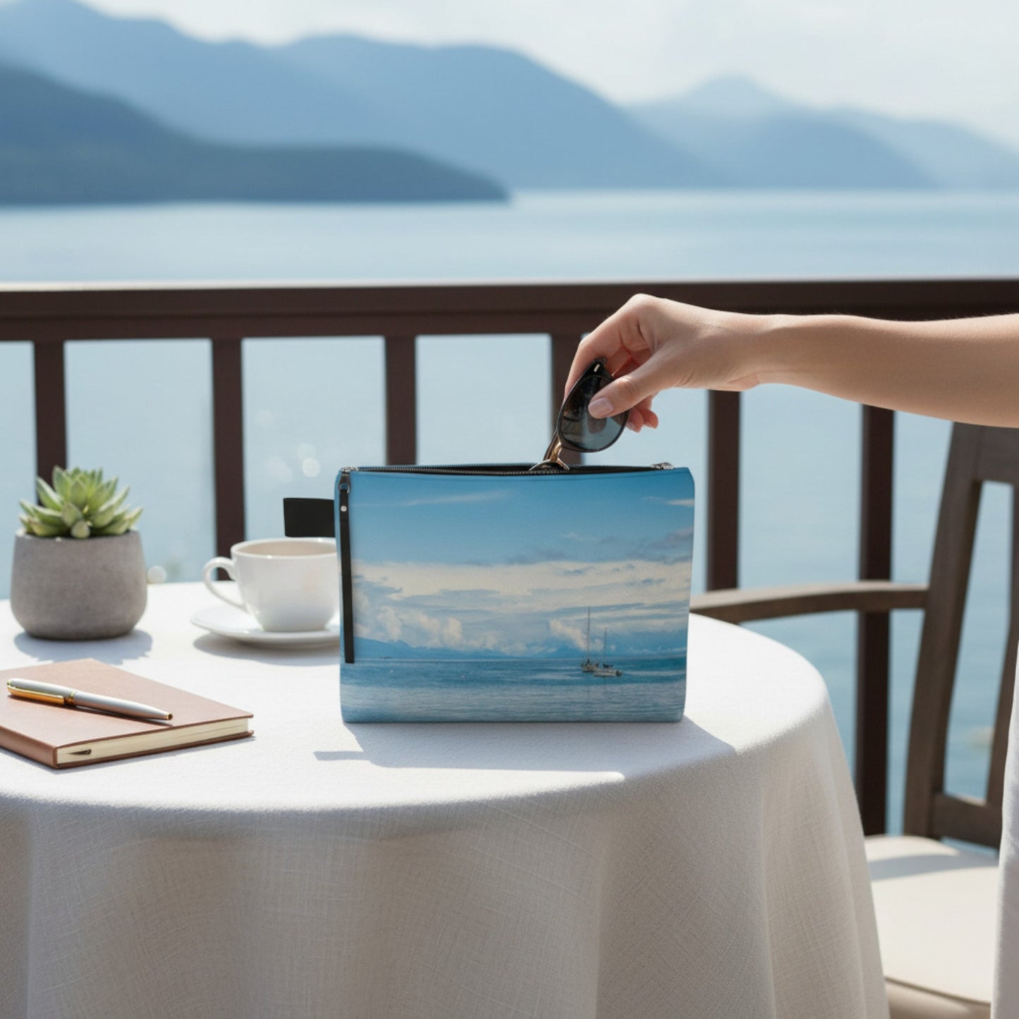 Hand reaching for a pouch with a scenic design on a table with a view of mountains and water.