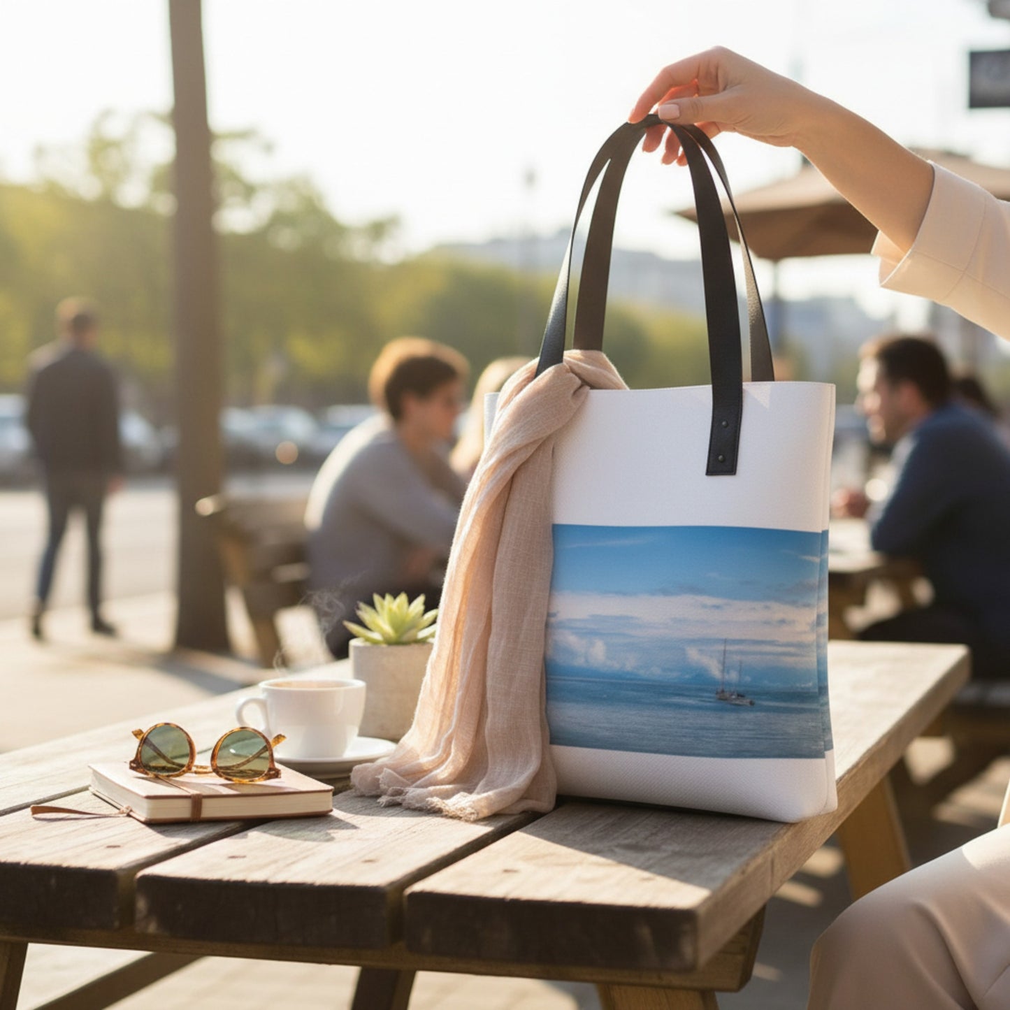 Person holding a tote bag with a beach design on a wooden table outdoors.