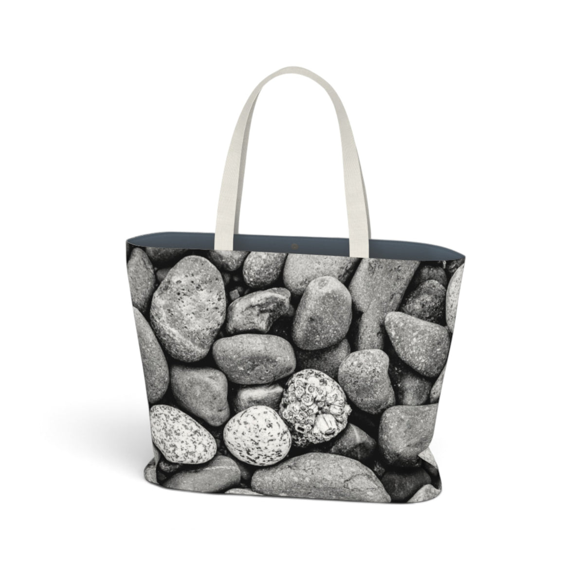 A tote bag featuring a black and white photograph of smooth river stones, with light beige fabric handles and a dark navy blue interior lining, shown against a plain white background.