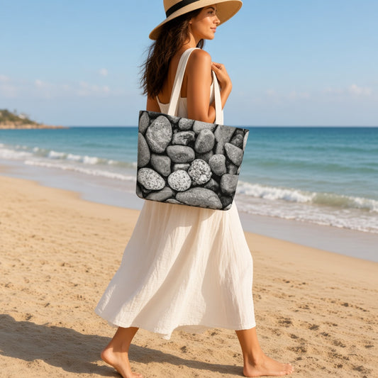 A woman walking on a sandy beach wearing a straw hat and a white dress, carrying a large tote bag with a black and white pebble pattern.