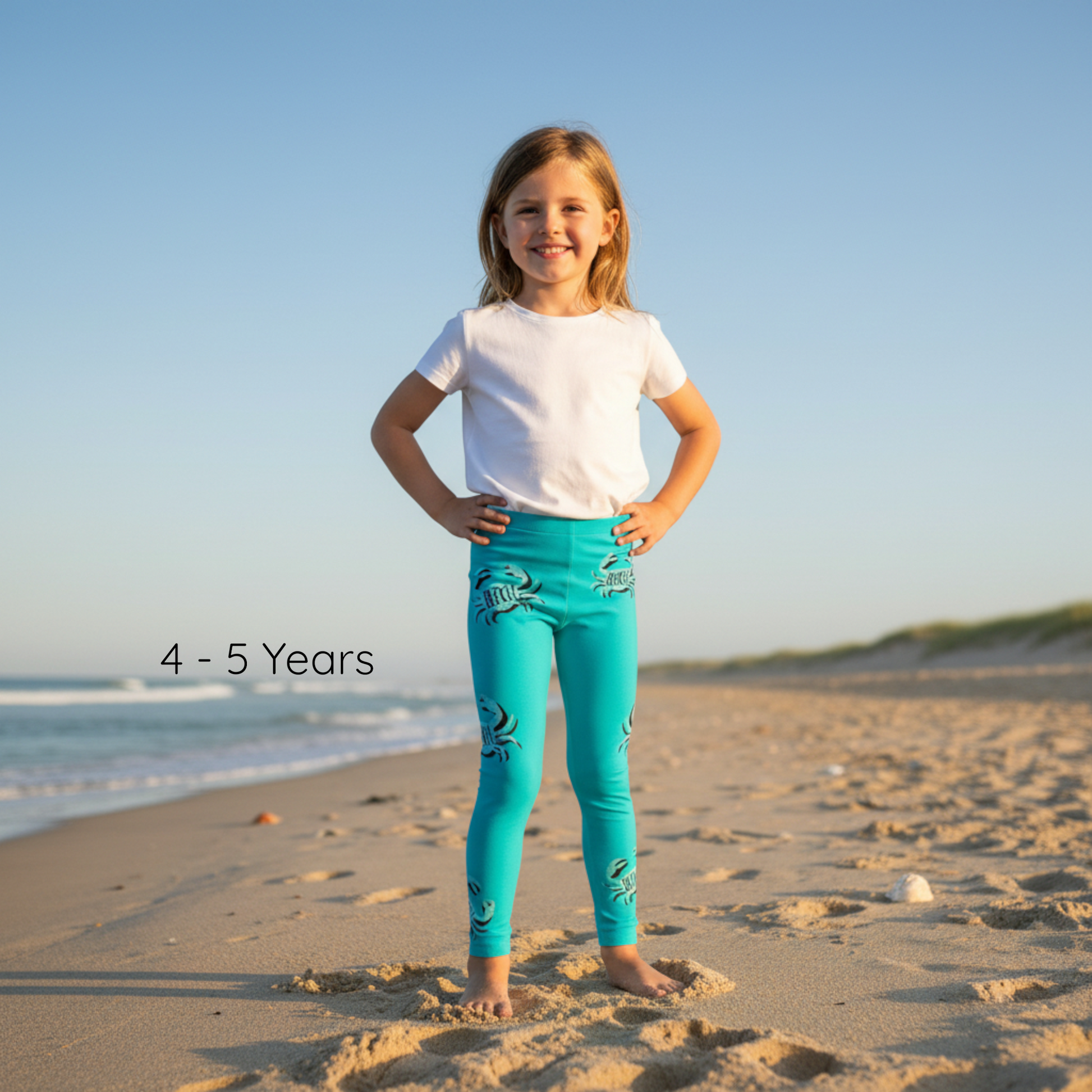 Young girl in turquoise leggings and white shirt standing on a sandy beach with ocean view.