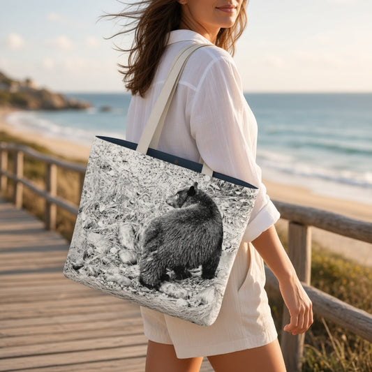 A woman on a wooden boardwalk by the beach carries a large tote bag printed with a black-and-white photo of a bear on rocks. The tote has cream shoulder straps and a dark navy top edge.