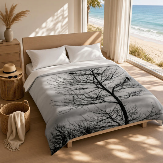 A bed dressed in a white duvet cover featuring a large black tree silhouette print, styled in a bright beach-side bedroom with a wooden headboard, bedside table, woven basket, and ocean view through large windows.