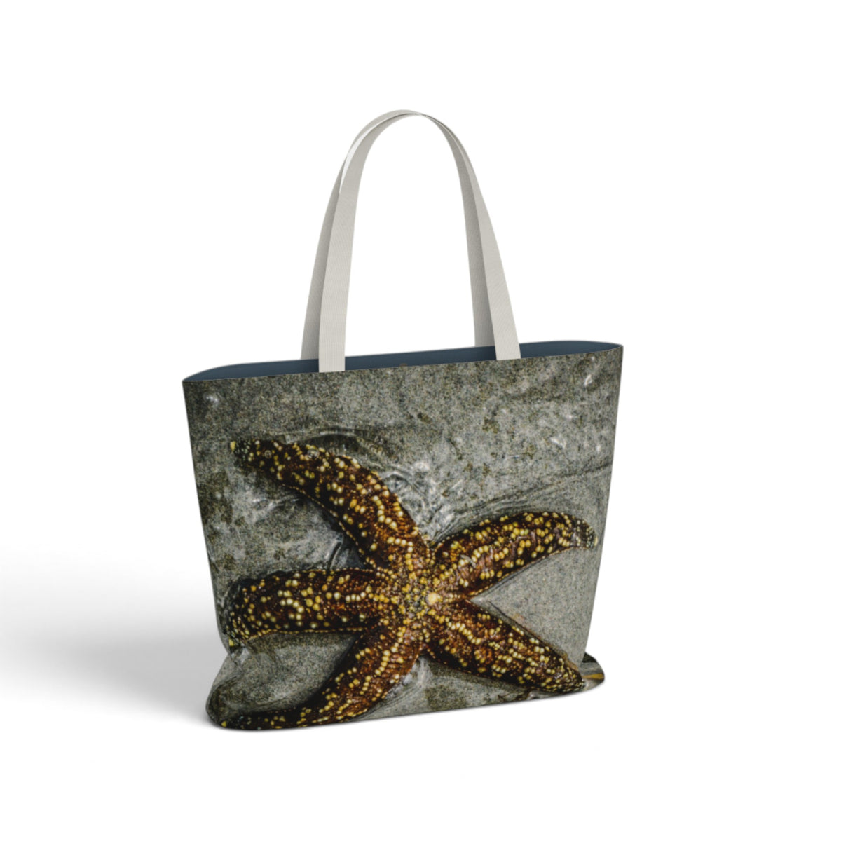 A tote bag featuring a real print of a brown and gold starfish on a gray stone-textured background, with light gray handles and a dark navy blue interior lining, shown on a white background.