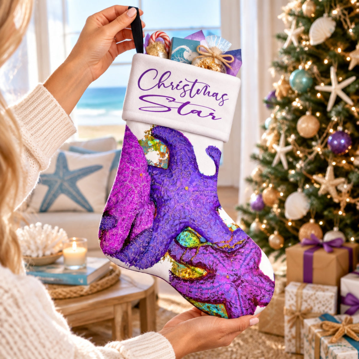 A person holds a Christmas stocking decorated with a purple and magenta marble pattern, gold glitter accents, and a white cuff that reads 'Christmas Star' in purple script. The stocking is filled with wrapped gifts and is shown in a cozy, decorated holiday home setting with a lit Christmas tree and beach decor in the background.
