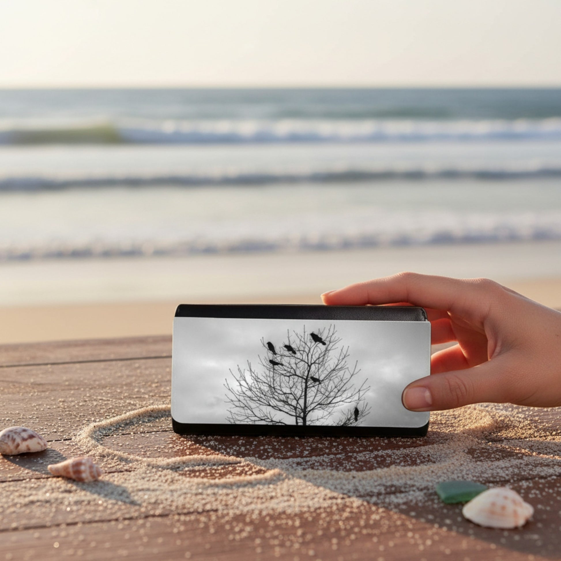 Hand holding a rectangular device with a tree and bird design on a beach.